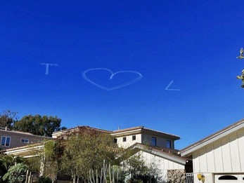 T heart L skywriting message in flowing cursive letters and a heart symbol above a residential neighborhood with luxury homes, created in collaboration with The Skywriters, showcasing precision skytyping, skywriting, aerial advertising, and high-impact thoughtful gestures, and creative proposal and memorial services. 