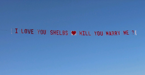 Custom airplane banner for a marriage proposal event announcing I Love You Shelbs followed by a red heart icon and Will You Marry Me, flying against a clear blue sky, created in collaboration with The Skywriters, showcasing professional aerial banner advertising, unique marriage proposal ideas, and high-impact aerial messaging for romantic celebrations.
