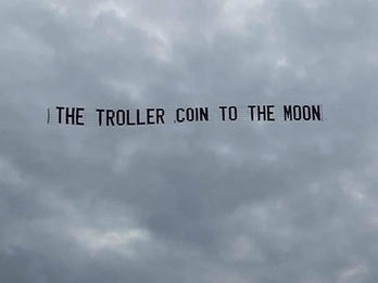 Sky banner featuring the message "THE TROLLER COIN TO THE MOON" in black block letters, flying against a dense, cloudy gray sky, created in collaboration with The Skywriters, showcasing precision aerial advertising and high-impact event marketing.
