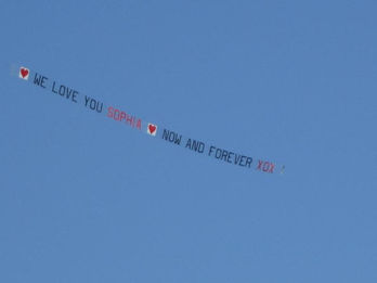 Sky banner featuring the message "heart WE LOVE YOU SOPHIA heart NOW AND FOREVER XOX" in a mix of black and red block letters, flying against a clear blue sky, created in collaboration with The Skywriters, showcasing precision aerial advertising, thoughtful sky messages, and creative airplane sky banners.