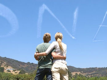 ONE skywriting message in flowing cloud letters in a clear blue sky above a couple embracing while overlooking a hilly landscape, created in collaboration with The Skywriters, showcasing precision skytyping, skywriting, aerial advertising, and high-impact event marketing, and creative proposal and thoughtful memorial ideas. 