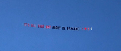 Custom airplane banner for a marriage proposal event saying Its All That Way Marry Me Pamcake, flying against a blue sky, created in collaboration with The Skywriters, showcasing professional aerial banner advertising, unique marriage proposal ideas, and high-impact aerial messaging for romantic celebrations.
