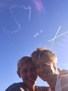 SIX skywriting message in flowing cloud letters above Ellen DeGeneres and Portia de Rossi in a sunny blue sky, created in collaboration with The Skywriters, showcasing precision skytyping, skywriting, aerial advertising, viral messaging opportunities, and creative wide area thoughtful gestures. 