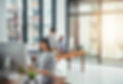 Mid range shot of woman at desk working on computer while colleagues chat behind her. 