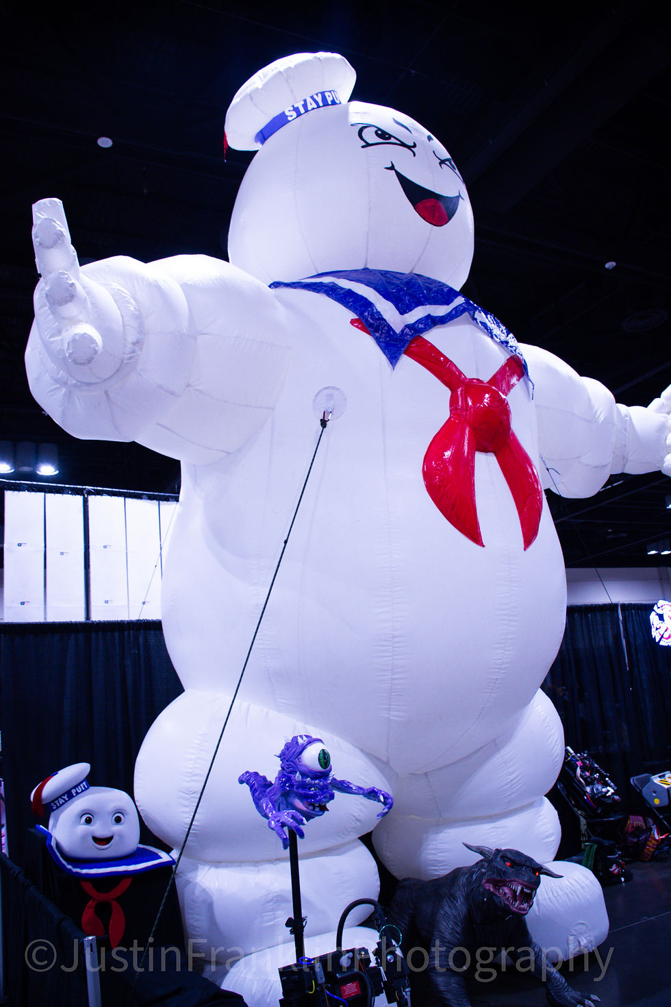 Inflatable white character with red scarf and blue collar, set against a dark indoor background. Nearby, a purple creature and wolf statue.