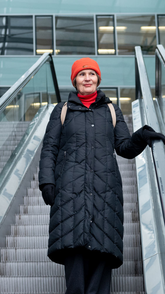 Gerda fährt eine Rolltreppe in der Mitte eines U-förmigen Gebäudes herunter. Sie trägt eine rote Mütze, einen roten Schal und einen langen schwarzen Mantel. 