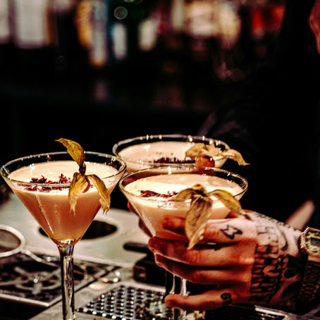 Tattooed hand holding three cocktail glasses garnished with physalis. Blurred background with colorful bottle bokeh, creating a vibrant mood.