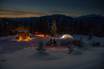 A wintery landscape with a snowcat, igloo lit up and snowmobiles. People hang out watching the sunset