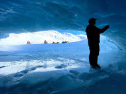 A man in an ice cave looks at the ice walls with a bright background of snow, snowmobiles and white snow