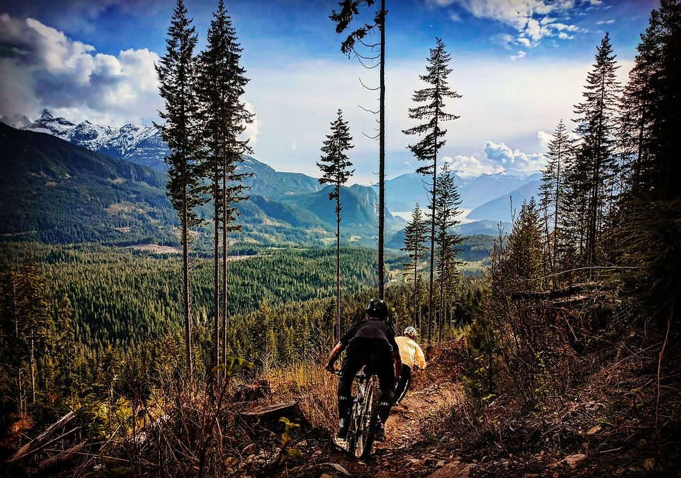 Two mountain bikers ride down a forested trail with snow-capped mountains in the background under a partly cloudy blue sky. Squamish.