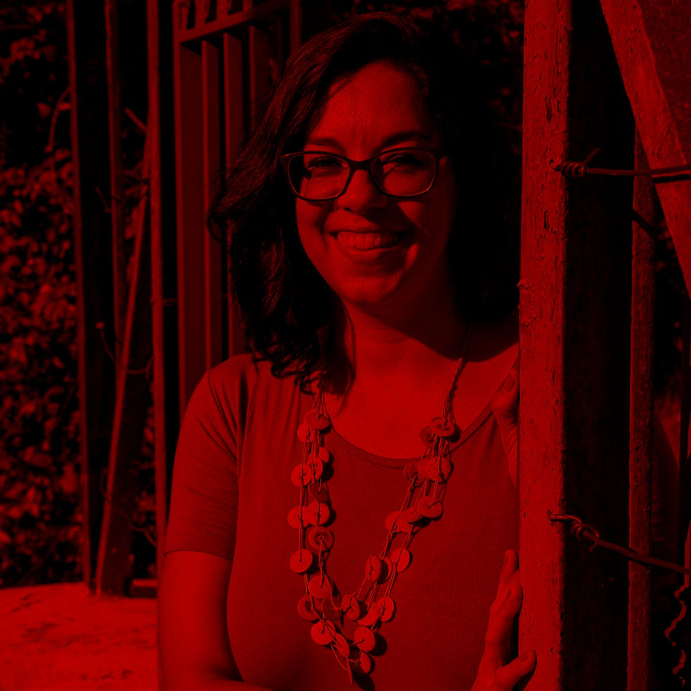 Mulher de cabelos escuros usa óculos, blusa laranja e colar artesanal, sorrindo em área externa ensolarada.