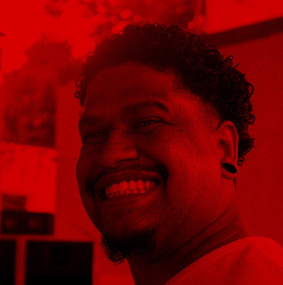 Rafael Rodrigues, homem negro de cabelo enrolado sorri amplamente, usa alargador e camiseta laranja clara.