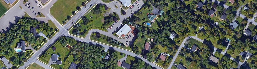 An aerial satellite view of a suburban neighborhood featuring tree-lined streets, residential houses, and several open green spaces. The image includes a school or community building with parking lots and sports fields, surrounded by dense greenery and roads connecting the area.