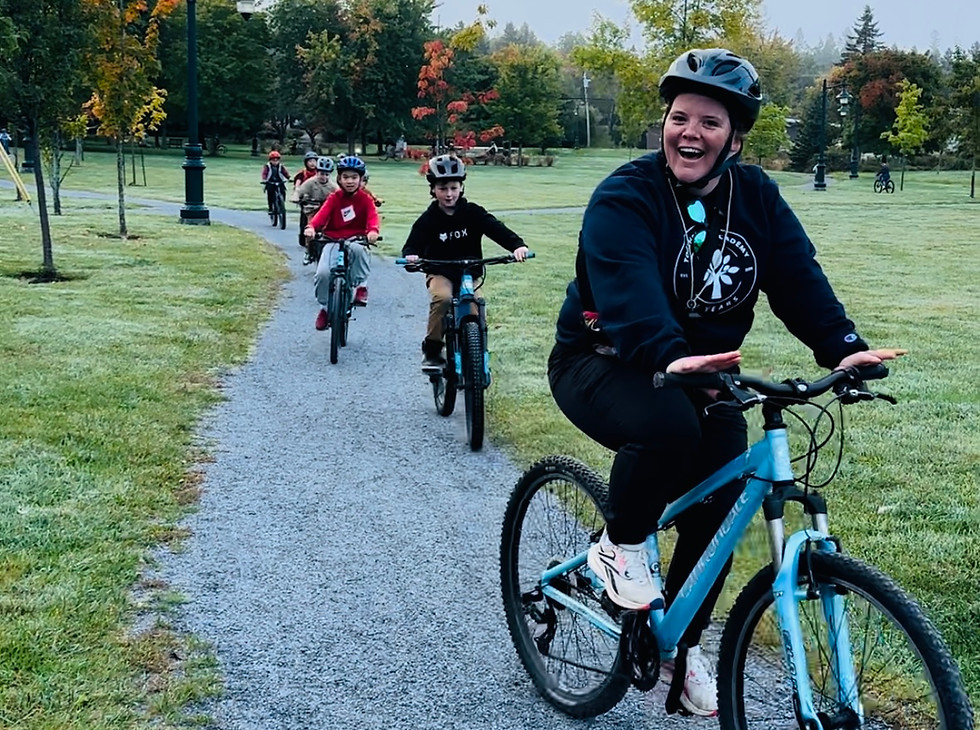 A group of children and an adult riding bicycles on a gravel path through a park. The adult, in the foreground, is smiling and wearing a navy hoodie and a helmet, leading the group. The park features green grass, trees with autumn colors, and a lamppost, creating a serene outdoor setting.