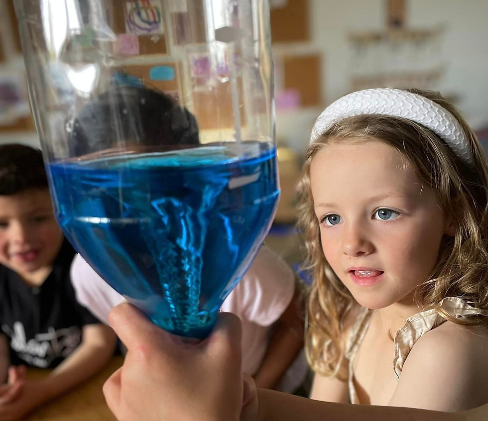 A group of young children observing a science experiment where a clear plastic bottle is filled with blue liquid, forming a swirling vortex. The focus is on a girl with blonde hair and a white headband, looking intently at the bottle being held by an adult. The background shows other children and a classroom setting.
