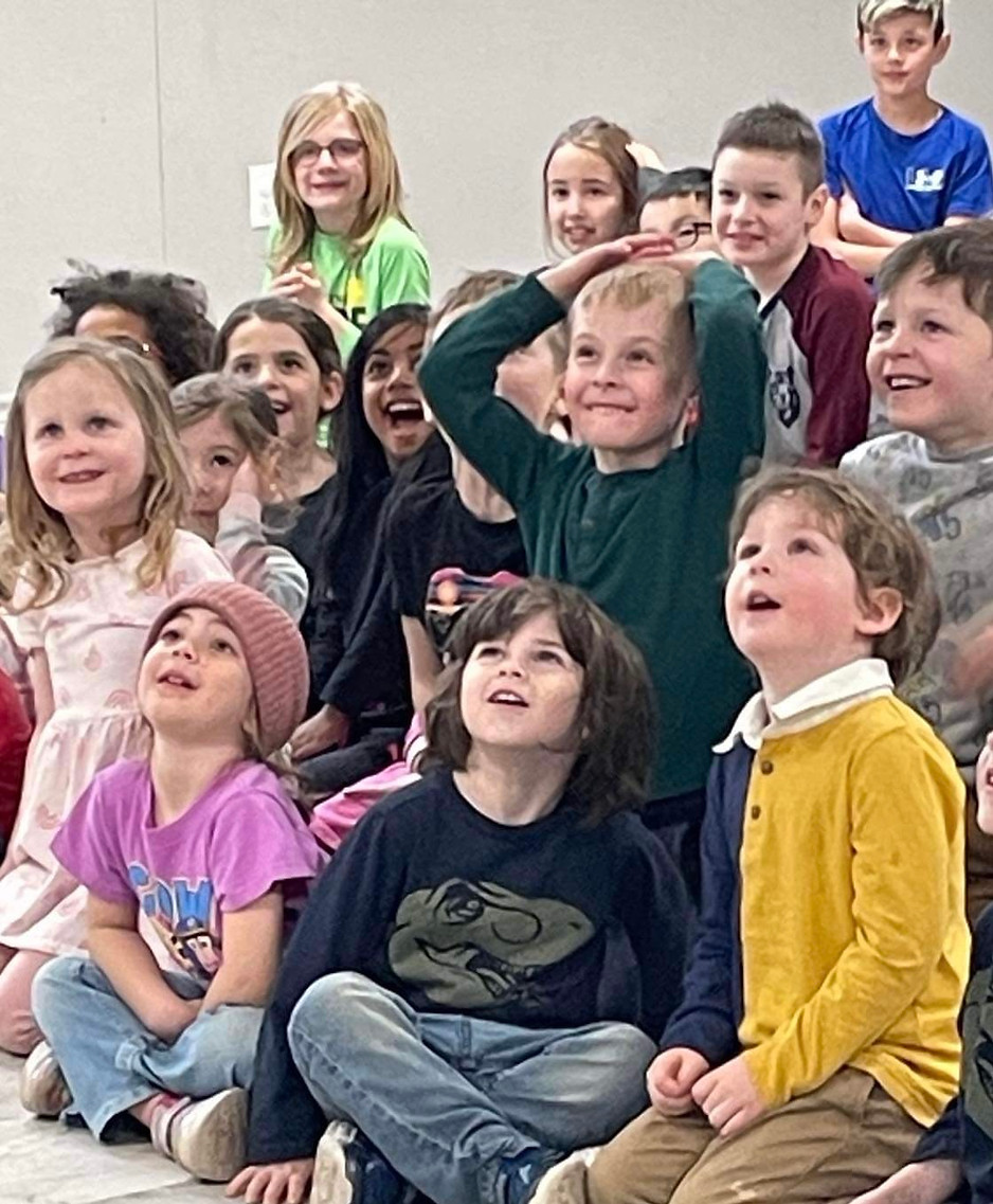 A group of young children sitting and standing in a classroom, all looking up with smiles and expressions of excitement and wonder. The children are dressed in colorful clothing, and the atmosphere appears joyful and engaging.