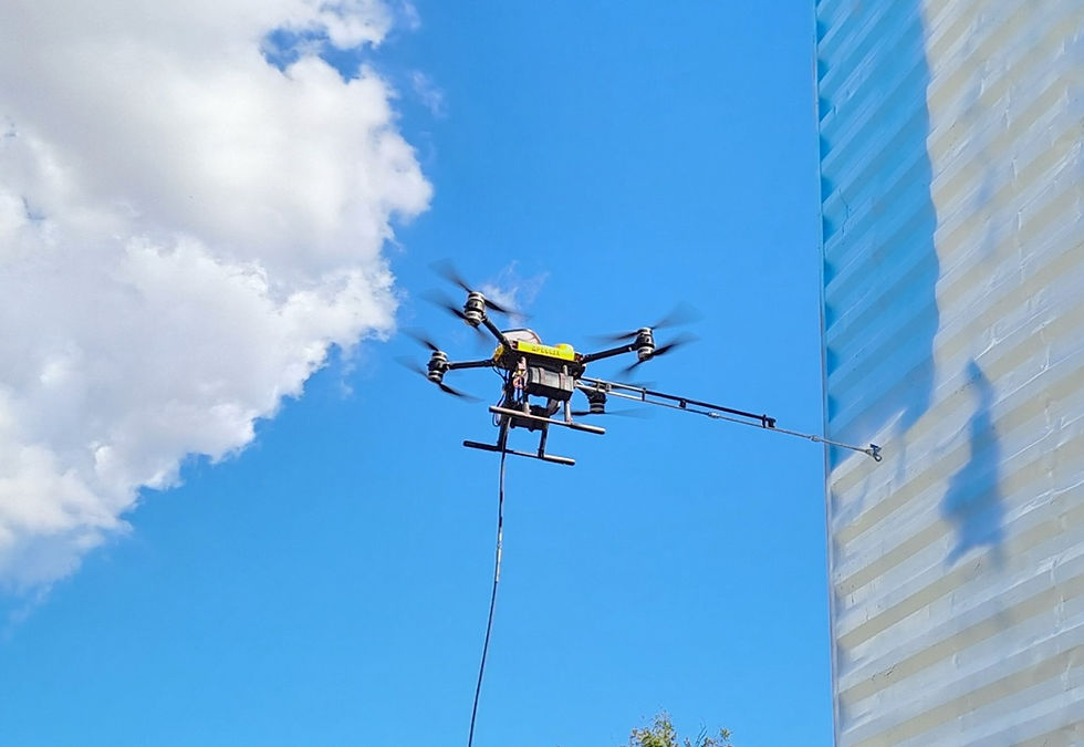 Drone-mounted cleaning system spraying the exterior wall of a tall building against a bright blue sky with clouds.