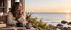A woman smiles while she looks out the window at the ocean and beach. There is a cup of coffee on the table in front of her.