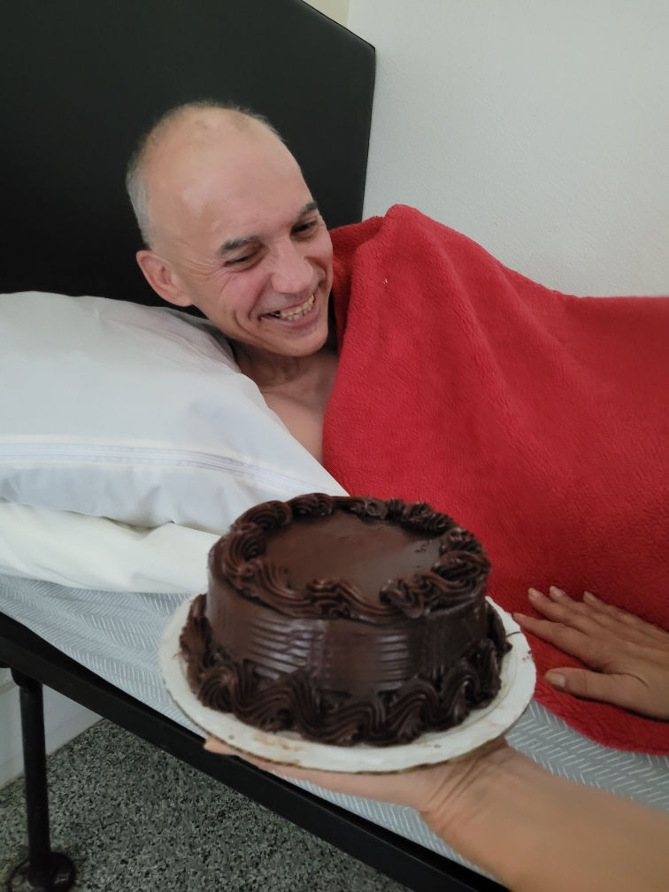 Joaquin, middle-aged, Hispanic man, lays in bed covered with fuzzy red blanket. His smile beams as he looks at a round chocolate cake being held before him.