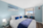 Bright bedroom with a large bed, blue accents, marina view, and cozy seating by the window. Coastal decor with ocean artwork and soft blue walls
