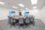 An octogon conference table with office chairs in an empty room. Two screens show a beach scene. Neutral tones, modern ambiance.
