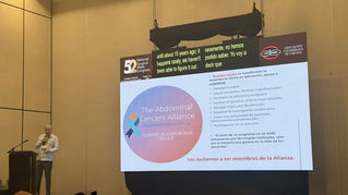 Dr. Sardi beside a conference screen showing The Abdominal Cancers Alliance slide with Spanish bullet points and logos.