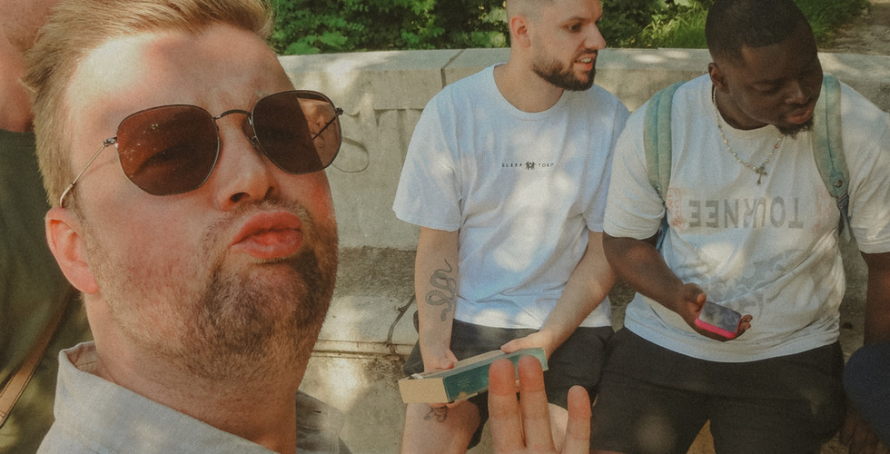 Mehrere vystem Teammitglieder machen ein lustiges Selfie im Englischen Garten. Eine Person steht nah an der Kamera, zwei weitere Teammitglieder sind im Hintergrund mit Spielunterlagen und Getränken zu sehen.