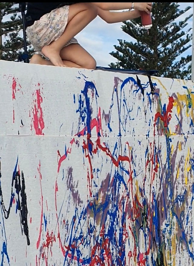 Large canvas over a wall cover with splashes on mutlic-oloured paint. Child sitting on top of the wall about the squeeze paint bottle.