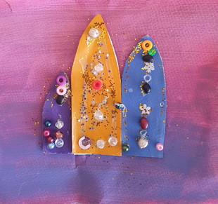 3 surfboards painted purple, orange and blue covered with gems and glitter stuck onto a purple pink painted background