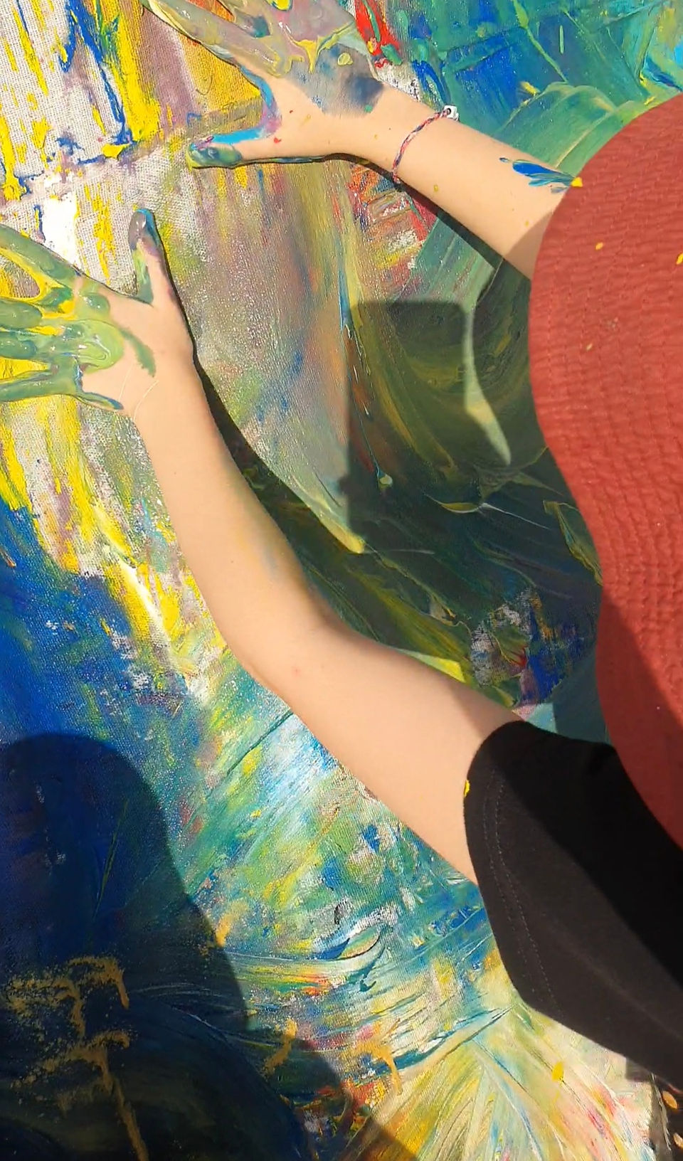 Child wearing an orange hat is using hand to smear green and yellow paint across a large canvas.