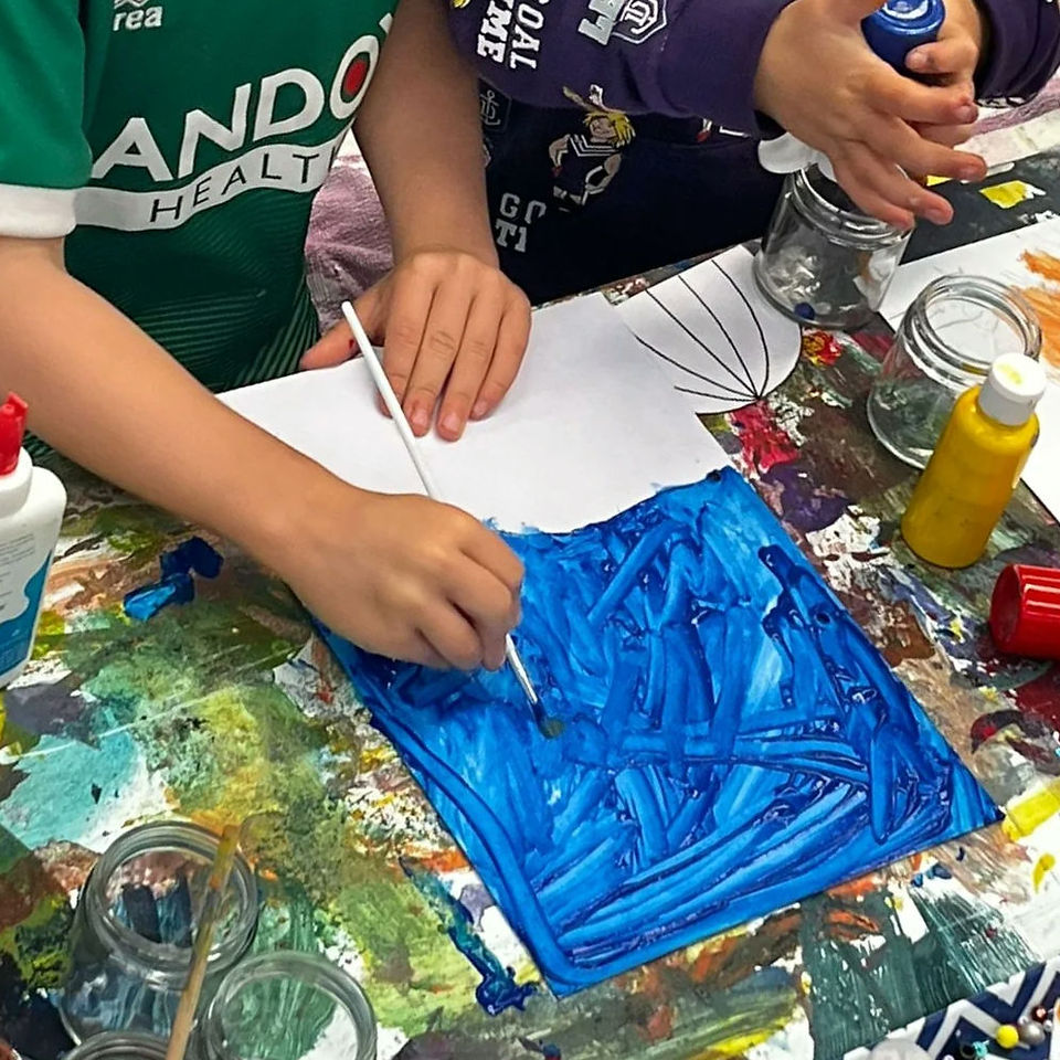 kids painting side by side. one painting onto blue canvas the other squeezing blue paint into a jar. 
