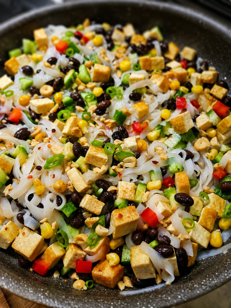 Colorful tofu and rice noodle stir-fry with black beans, corn, cucumbers, peppers, scallions, and nuts in a skillet.