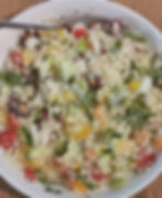 A colorful couscous salad with tomatoes, cucumbers, peppers, onion and greens in a white bowl. A fork rests inside, set on a wooden table.