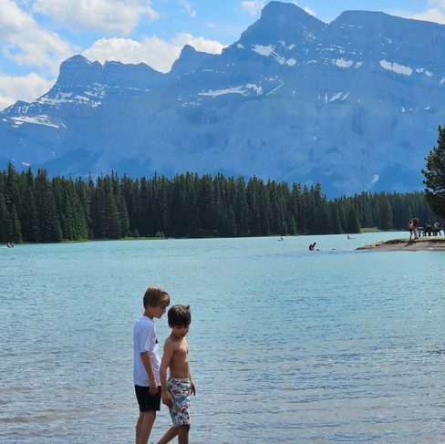 two boys walking near a lake surrounded by mountains and trees
