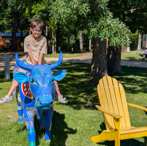 a kid on a blue ceramic bull on a grassy area near a yellow chair, in a campground
