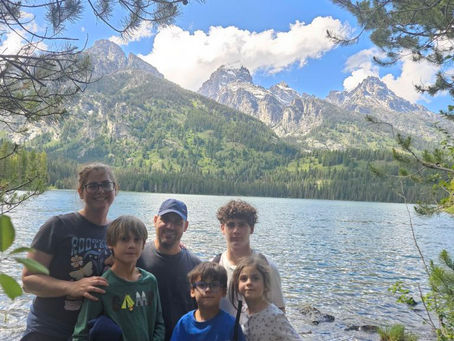 Smiling family of five stands by a mountain lake, framed by pine branches, with snowy peaks and blue sky behind them.