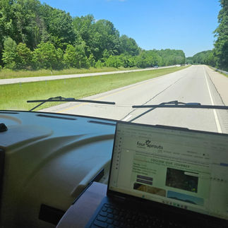 Inside an RV on a sunny highway, a laptop shows the Four Sprouts website while green trees line the road.