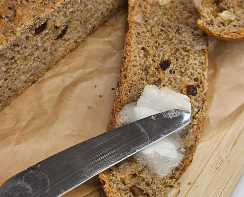 Slice of nutty herbal spelt bread spread with vegan butter, with whole loaf in the background on parchment paper.