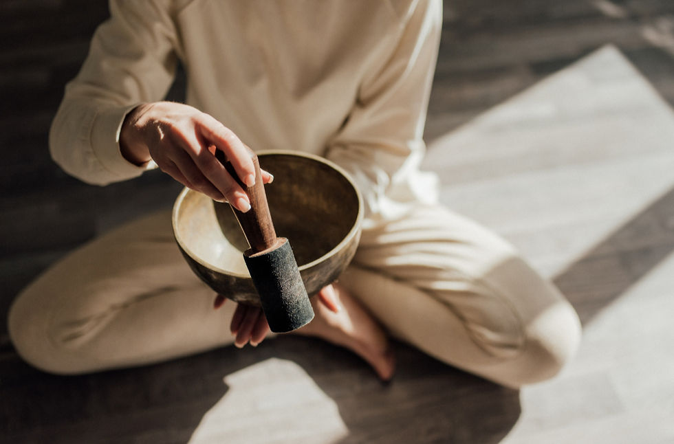 Person in light clothing sitting on the floor, playing a singing bowl with a mallet. Sunlight casts patterns on wooden floor, serene mood.