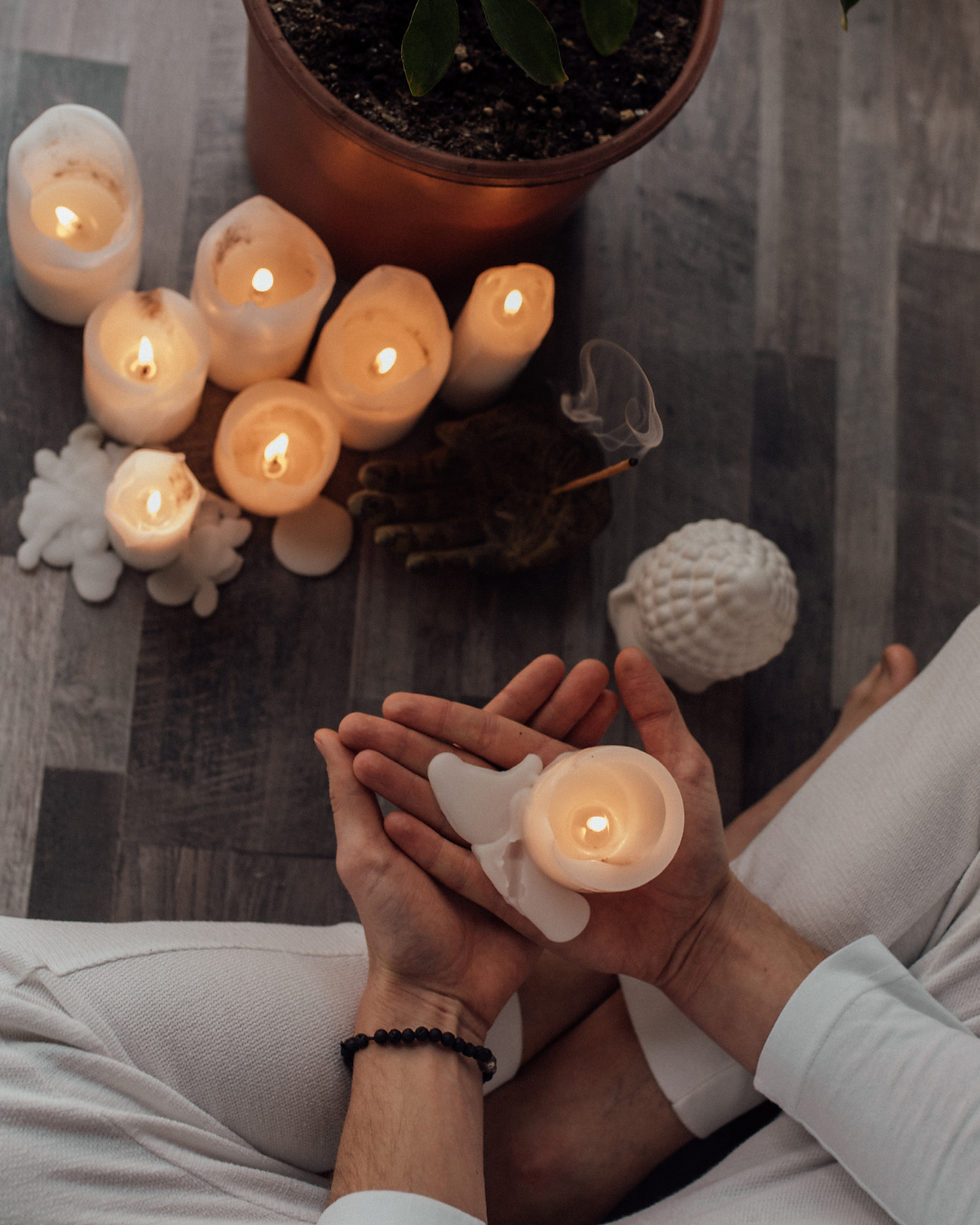 Rituals for the 12 Houses. Hands holding a lit candle with angel wings, surrounded by other candles and a potted plant on a wooden floor, creating a calm atmosphere.