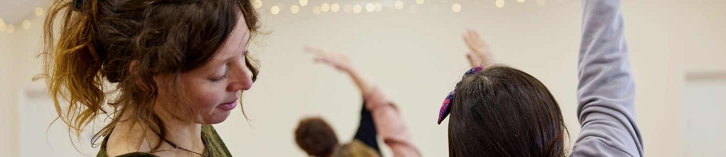 Side view of a woman's face, she is looking at a group of women reaching the'r arms upwards in a yoga pose, pale background and faint fairy lights on the cieling