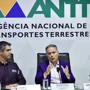 Três homens em conferência na ANTT. O central fala gesticulando. Fundo com logotipo e texto "Agência Nacional de Transportes Terrestres".