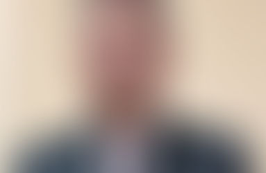 Homem de terno preto e gravata roxa em pé, mãos cruzadas. Fundo bege. Expressão séria. Ambiente interno.