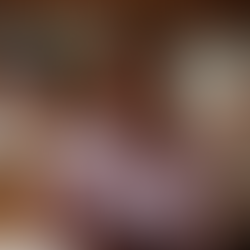 Mulher de blusa lilás gesticula animadamente, sentada ao lado de taça de vinho em ambiente interno. No fundo, bandeira colorida.