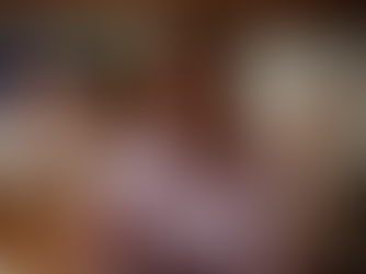 Mulher de blusa lilás gesticula animadamente, sentada ao lado de taça de vinho em ambiente interno. No fundo, bandeira colorida.