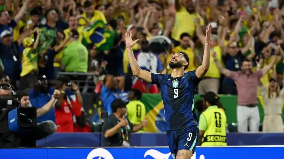 Jogador do Brasil, camisa 9 azul, ergue os braços em comemoração diante de torcida com bandeiras do Brasil no estádio.