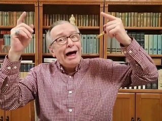 Homem de óculos e camisa xadrez gesticula animadamente com as mãos para cima. Fundo com estante de livros, expressão alegre.