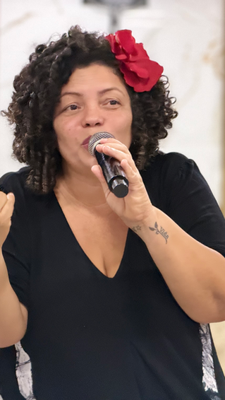 Mulher com flor vermelha no cabelo fala ao microfone em uma reunião. Fundo com bandeira colorida e números 13 visíveis. Ambiente sério.
