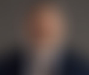 Homem de óculos, vestindo terno azul e gravata xadrez, olha seriamente para a câmera. Fundo cinza, imagem formal e profissional.