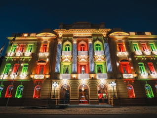 Fachada de prédio histórico à noite, iluminada em verde, vermelho e dourado, com palmeiras e céu escuro ao fundo.